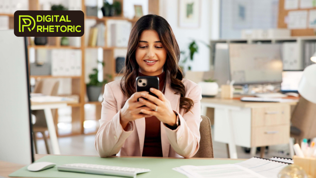 A smiling woman in a pink blazer uses her smartphone at an office desk. The lime green "Digital Rhetoric" logo is visible on the desk and background office surfaces